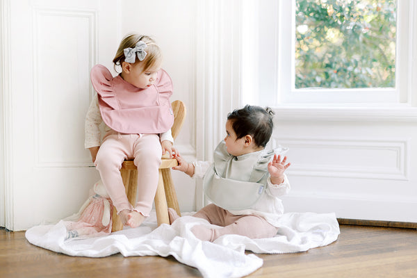 a baby girl sitting wearing a ruffle angel baby bib and toddler girl on stool wearing a dark pink ruffle angel bib