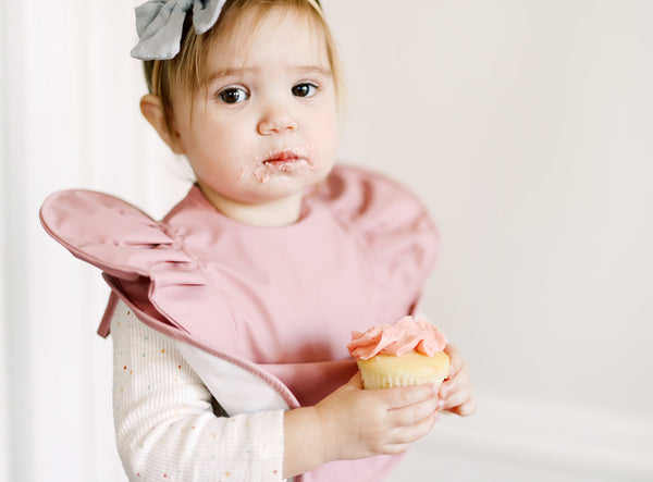 toddler girl wearing a pink ruffle baby bib holding a cupcake